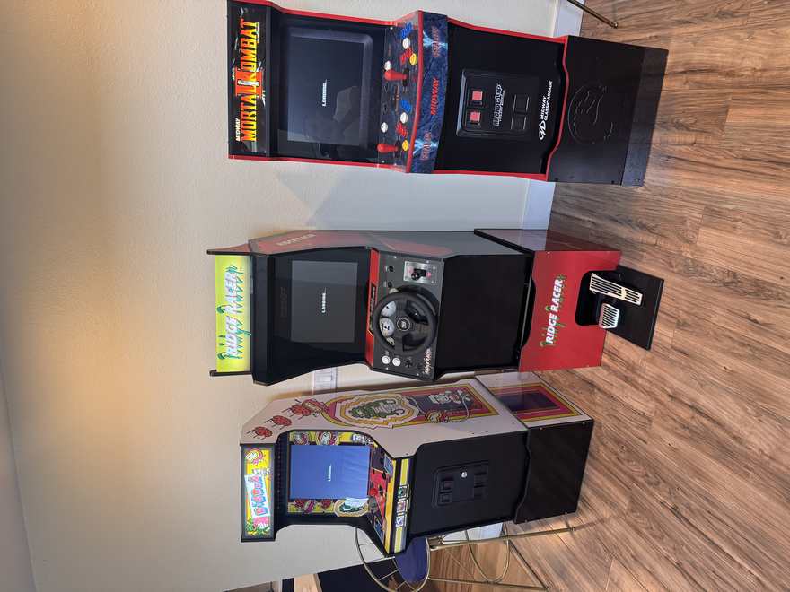 Three upright arcade cabinets against a wall: Dig Dug, Ridge Racer with a steering wheel and floor pedals, and Mortal Kombat II
