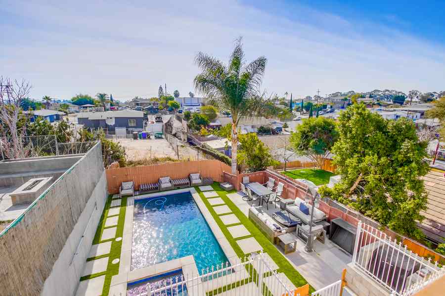 The back yard from above: a rectangular pool and raised spa inside a white iron fence, lounge chairs and a palm along one side, with the neighbourhood and hills beyond